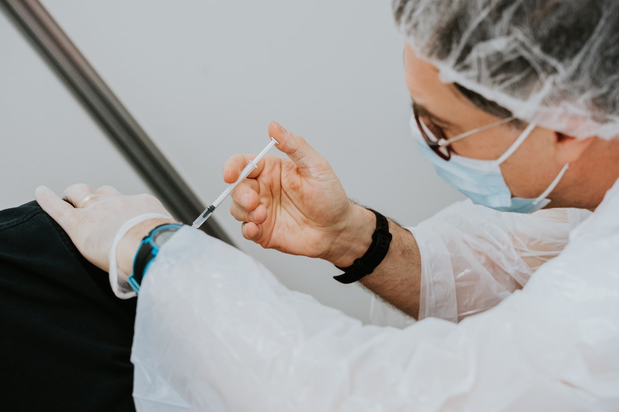 Image representing work related to clinical trials shows a masked healthcare professional wearing a white coat and gloves, performing an injection on a patient